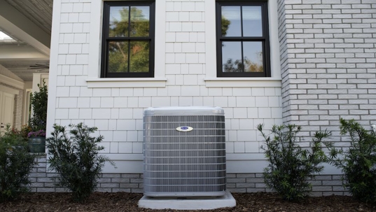 An outdoor HVAC unit sits in front of a white house with two windows.