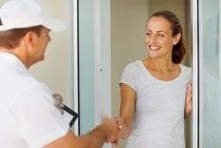 A person wearing a white hat and shirt is shaking hands with a smiling woman at her front door.