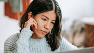 A young woman in a light blue sweater looks pensively at her screen.