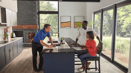 A man in a blue shirt presents documents to a couple in a modern kitchen.