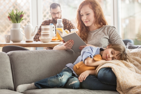 Family scene: a parent reads to a sleeping child; another parent sits at a table.