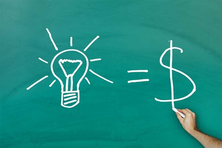 A hand is drawing a dollar sign on a chalkboard, with a light bulb and equals sign already on the board.