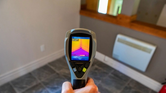 A hand holds a thermal imaging camera, pointed at a wall with an attached heater.